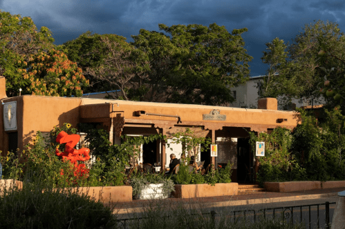 A charming adobe-style restaurant surrounded by greenery, with a cloudy sky in the background.